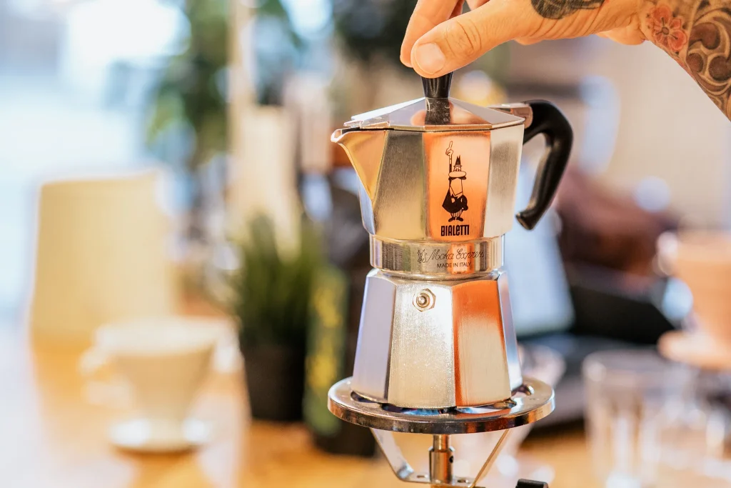 Tradycyjna, srebrna kawiarka aluminiowa Bialetti Moka Express trzymana za uchwyt nad palnikiem w kawiarni lub kuchni.