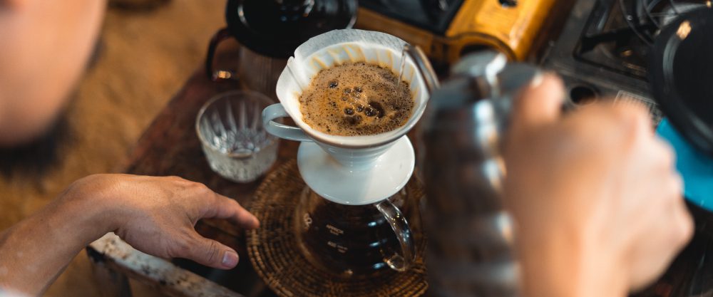 Przygotowanie kawy specialty metodą przelewową. Widok z góry na ceramiczny dripper V60 z papierowym filtrem, do którego baristy nalewa wodę ze specjalnego czajnika.